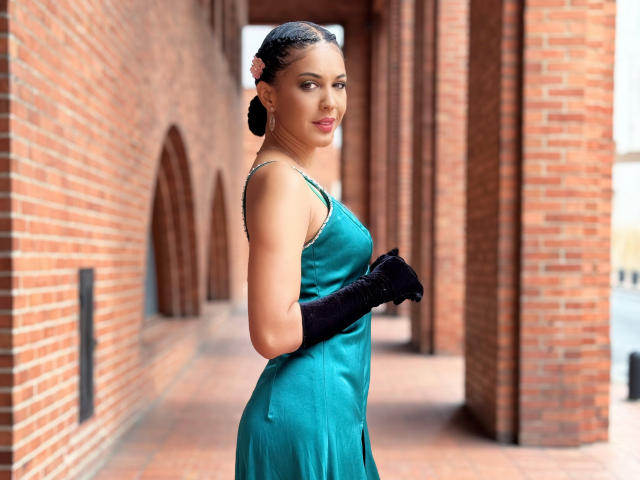 JoannaCool with dark skin, braided hair, teal dress, black gloves, looking over shoulder, brick hallway, small breasts, ass slightly visible, confident smile, pink lipstick, elegant pose.