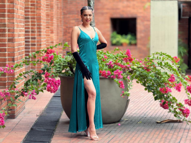JoannaCool stands in a teal dress with a high slit, black gloves, and heels, showing her ass with a leg raised, next to pink flowers in a large pot.