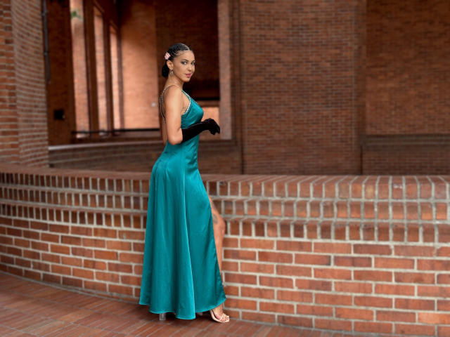 JoannaCool stands in a teal ball gown, black gloves, and sandals, holding a black fan, with her ass slightly visible, against a brick wall, looking over her shoulder.