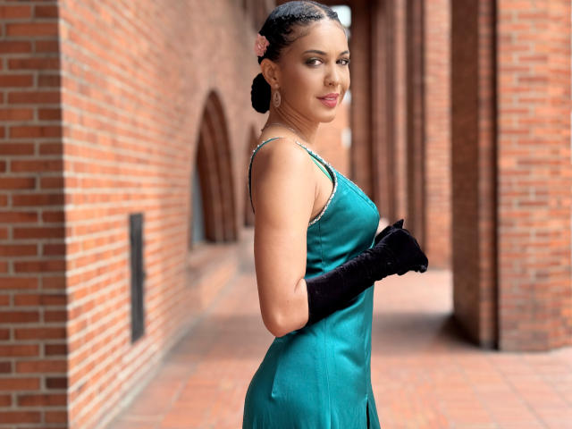 JoannaCool with black gloves, teal dress, and pink hairpin, standing in a brick hallway, showing off her curvy ass and small tits. She's looking at the camera with a smile.