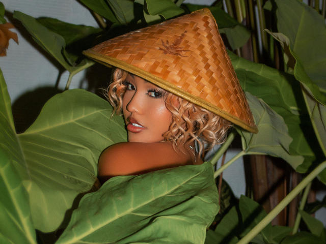 VictoriaXLi with curly blonde hair, wearing a bamboo hat, has her ass and shoulders covered by large green leaves, looking seductively at the camera.
