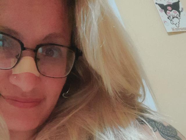 RusherRed with blonde hair, glasses, nose piercing, and a slight smile, showing a tattoo on their shoulder, and a cute, fluffy nose in a selfie.