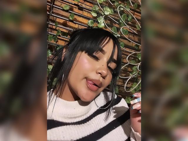 KarinaBlake with black hair, pink lips, and a nose ring, sticking out her tongue, wearing a black and white striped sweater, against a wooden and green leafed background.