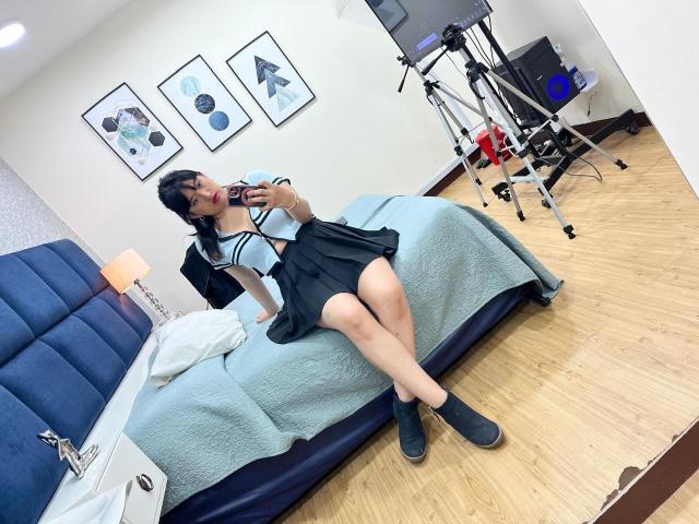 CarolineHarpper sitting on a bed, wearing a schoolgirl outfit with a blue skirt, white shirt, and black socks, holding a phone, her ass exposed, in a bedroom.