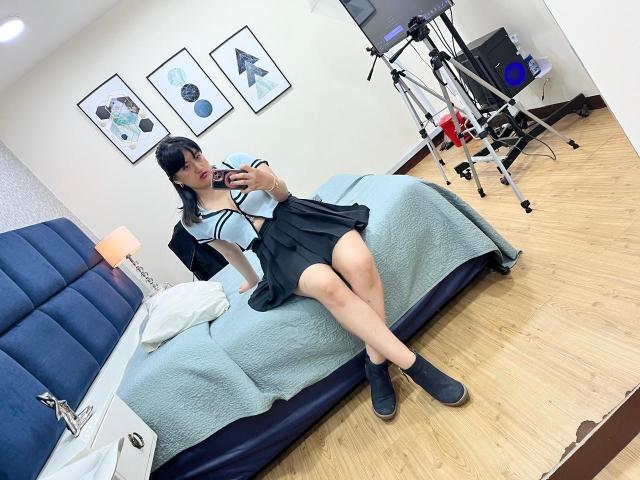 CarolineHarpper with black hair, wearing a blue and black schoolgirl outfit, sitting on a bed, taking a mirror selfie, showing her tits and ass, with a tripod in the background.