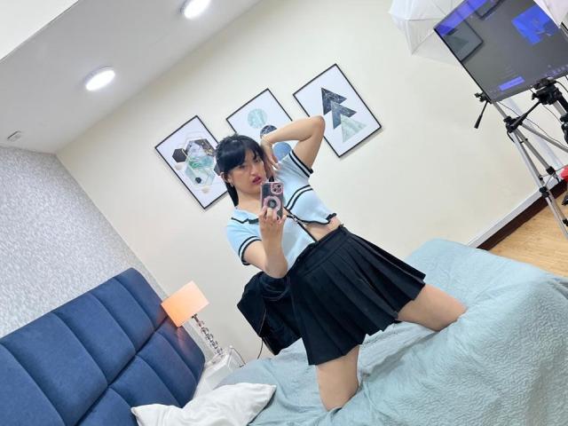 CarolineHarpper, kneeling on a bed, wearing a blue sailor top, black skirt, and red lipstick, taking a selfie with her phone, showing her small tits and ass.
