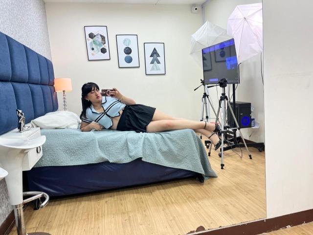 CarolineHarpper is lying on a bed, wearing a blue top and black skirt, taking a selfie with her phone, legs crossed, in a room with photography equipment.