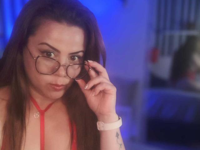 AstridBayon with long brown hair, wearing red strappy top, adjusting glasses, looking down with pouty lips, pink wristwatch, tattoo on wrist, blue lighting, soft focus.