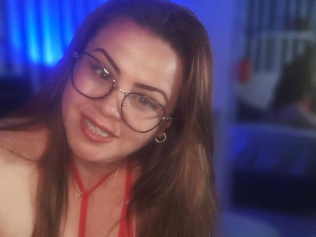 AstridBayon with glasses, brown hair, red halter top, smiling, showing teeth, blue background, hoop earrings, slightly blurry, cute, sexy, smiling directly at the camera.