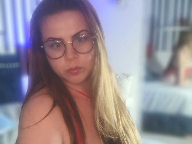 Astrid Bayon, con lunghi capelli castani, che indossa occhiali, una collana rossa e che guarda la fotocamera, mostrando il seno, in un ambiente sfocato che sembra una camera da letto..
