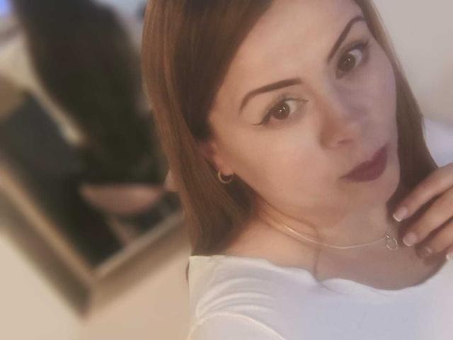 AstridBayon ha la pelle chiara, gli occhi marroni e i capelli lisci castani, indossa una maglietta bianca, rossetto scuro e una delicata collana, con un orecchino a cerchio dorato. La sua mano tocca il collo..