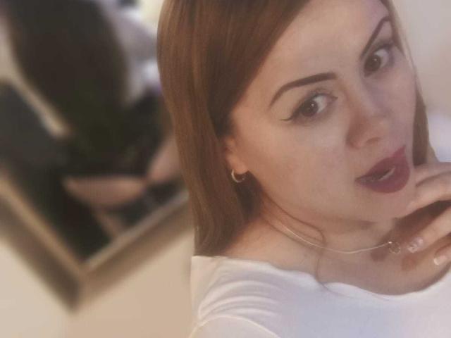 Astrid Bayon ha la pelle chiara, i capelli castani e l'eyeliner scuro. Indossa una maglietta bianca, rossetto rosso e un anello dorato. Le sue dita sono vicine alla bocca leggermente aperta..