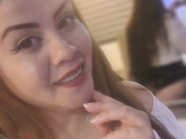 Astrid Bayon, con piel clara, cabello rojo y delineador de ojos, sonriendo, mostrando sus dientes, tocándose la barbilla con una mano, mostrando esmalte de uñas blanco, en una foto borrosa..