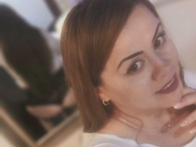 Astrid Bayon ha i capelli castano chiaro, le sopracciglia scure ed è sorridente, con il rossetto rosso. Indossa una maglietta bianca, piccoli orecchini a cerchio e una collana sottile. Il suo riflesso è visibile nello specchio..