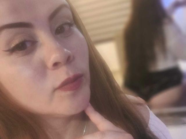 Astrid Bayon, con lunghi capelli castani, che indossa l'eyeliner, si tocca il mento, guarda la fotocamera, mostrando un riflesso della sua schiena e della biancheria intima nera nello specchio..