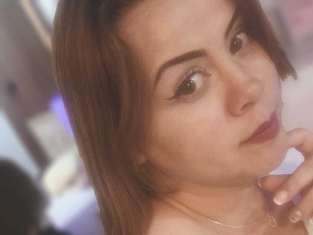 Astrid Bayon tiene la piel clara, ojos marrones y cabello castaño, con una ligera sonrisa y un dedo tocándose la barbilla. Lleva puesto un collar dorado..