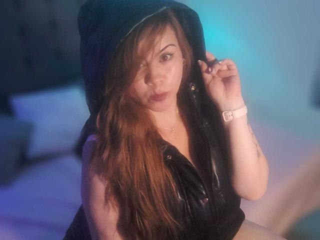 Astrid Bayon, con su largo cabello rojo, lleva una capucha de cuero negro que se está subiendo, tiene un aspecto atractivo, piel clara y un reloj rosa, y está sentada en una cama..