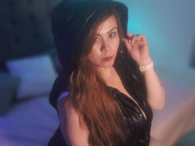 Astrid Bayon, com longos cabelos castanhos, vestindo um vestido de couro preto com capuz, que revela levemente seus seios, iluminação azul e roxa, olhando para o lado, tocando o cabelo..
