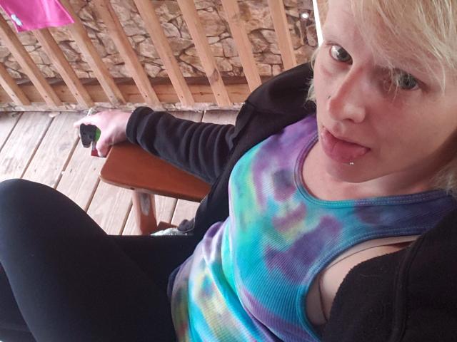 KaszyMarie with blonde hair, green eyes, and a lip piercing, wearing a tie-dye top, black jacket, and black pants, holding a phone, sitting on a wooden bench.