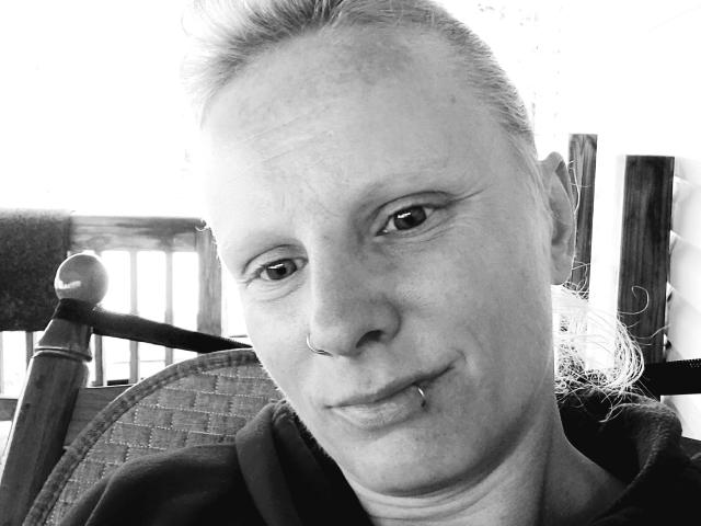 Black and white selfie of KaszyMarie with a nose ring and lip piercing, slightly squinting eyes, and a small smile, wearing a dark hoodie, and tied-back hair.