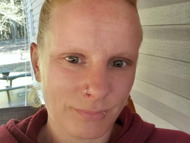 KaszyMarie has a nose ring and a lip piercing, with light skin, green eyes, and blonde hair in a ponytail, wearing a red hoodie.