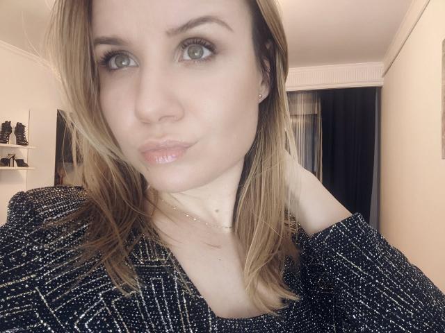 HeartlessQueenn met volle lippen, groene ogen en blond haar, draagt een zwarte, glinsterende trui waarbij haar schouder te zien is. Ze draagt een dunne ketting en kijkt iets opzij..