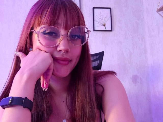 LiaMonroelu with long brown hair, glasses, nose ring, and pink lipstick, resting chin on hand, wearing black smartwatch, smiling. Purple light. Boobs not visible.