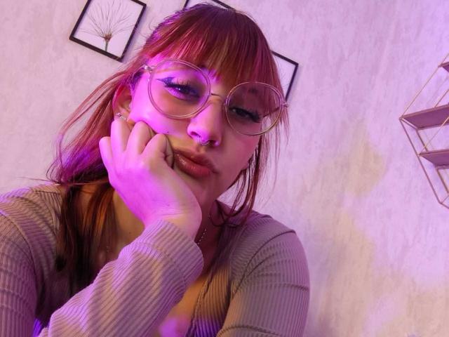 LiaMonroelu with pink-tinted hair, glasses, and nose ring, holding her face, wearing a ribbed gray top, looking down, purple light on her face.