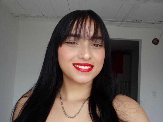 MaraLewis with black hair, red lipstick, and a nose ring, smiling at the camera, wearing a chain necklace, has red lipstick on her shiny teeth.