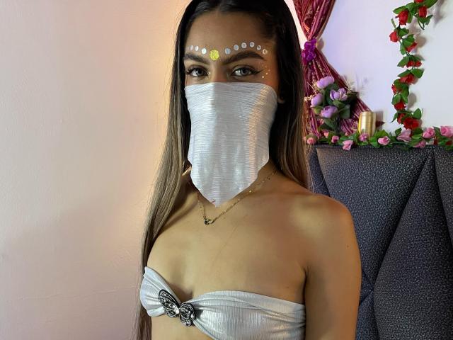 DalilaAliX with long brown hair, wearing a silver mask, white strapless top showing small tits, face paint with yellow dots, standing in a room with floral decor.