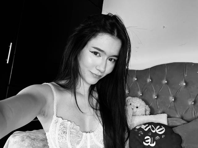 Black and white selfie of DeisyWest with long dark hair, wearing a lace top, showing her small tits, and a cute smile, with a stuffed teddy bear in background.