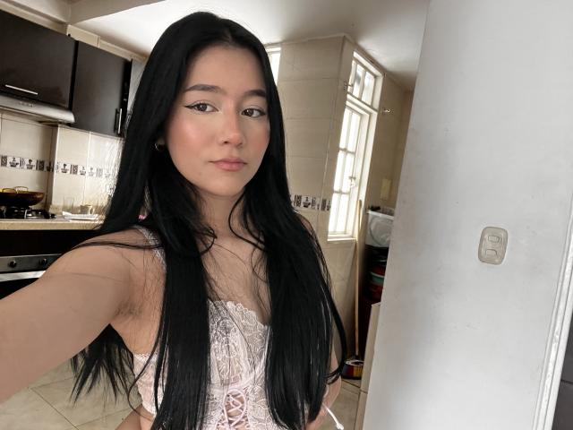 DeisyWest with long black hair, wearing a pink lace corset, showing her small tits, standing in a kitchen, taking a selfie.