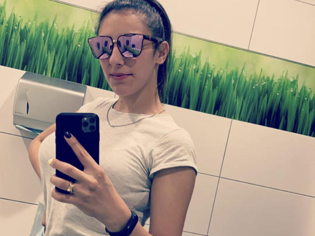 MelisseX with glasses, tied-back brown hair, white tee, holding black phone, showing small tits, gray background, ring on left hand, wrist bracelet, subtle smile, slightly visible nose piercing.