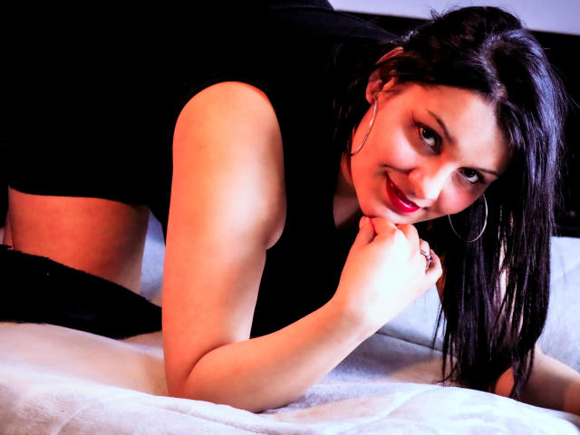 ScarlettMancini, with red lipstick and black dress, is on all fours on a bed, smiling at the camera, her hand near her mouth, showing her tits and ass.