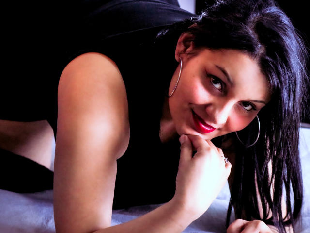 ScarlettMancini with dark hair, red lipstick, hoop earrings, black sleeveless top, lying on bed, smiling, resting chin on hand, visible bare shoulders, soft lighting.