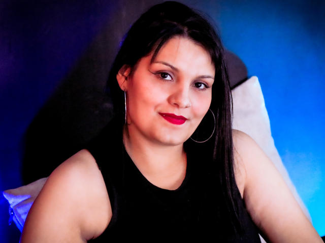 ScarlettMancini with dark straight hair, red lipstick, black sleeveless top, and hoop earrings, smiling softly at the camera, showing her cute, chubby face and small tits.
