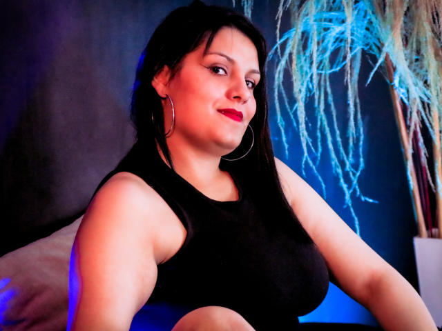 ScarlettMancini with dark hair, red lipstick, black sleeveless top, hoop earrings, smiling, blue and pink light, holding phone, visible breasts, slightly turned, puffy pillow in background.