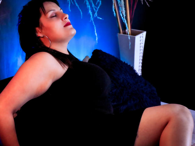 ScarlettMancini with her eyes closed, black dress, red lipstick, and hoop earrings, sitting back, arms behind, showing her ass, on a black couch, against a blue light.