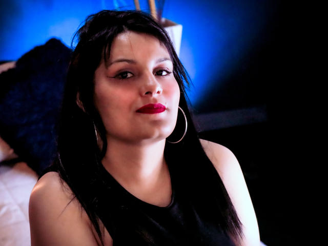 ScarlettMancini with dark hair, red lipstick, hoop earrings, and black sleeveless top, looking slightly to the side, in a dimly lit, blue-toned room.