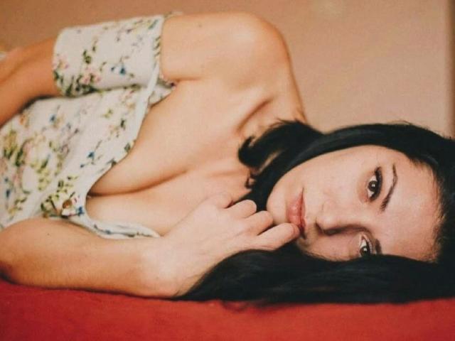 LolaBlaze-hot with dark hair, lying on red bed, wearing floral dress, exposing small tits, looking at camera, hand near chin, soft brown eyes, gentle expression.