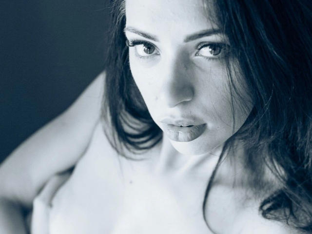 LolaBlaze-hot with serious eyes, dark wet lips, and messy dark hair, looking up. She's topless, showing small tits, with a soft, gray-blue, slightly blurred photograph style.