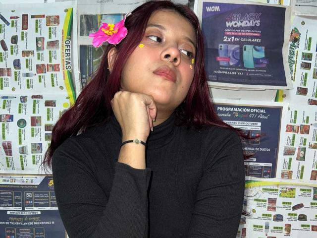 CoralineHale with dark red hair, a pink flower in her hair, and yellow dot makeup, wearing a black long-sleeve shirt, resting her chin on her hand, looking bored.
