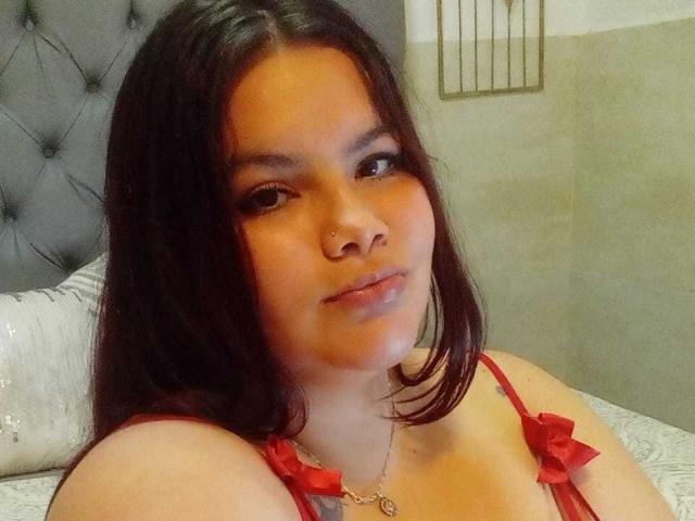 CelesteSoler with dark hair, wearing red lingerie with bow ties, nose piercing, and a necklace, sitting on a bed, looking slightly to the side, with a neutral expression.