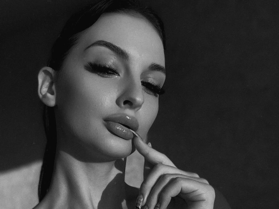 KessyX with glossy lips, dark eyebrows, and long eyelashes, biting a finger with manicured nails, looking down, in a black-and-white photo. Her wet hair is pulled back.
