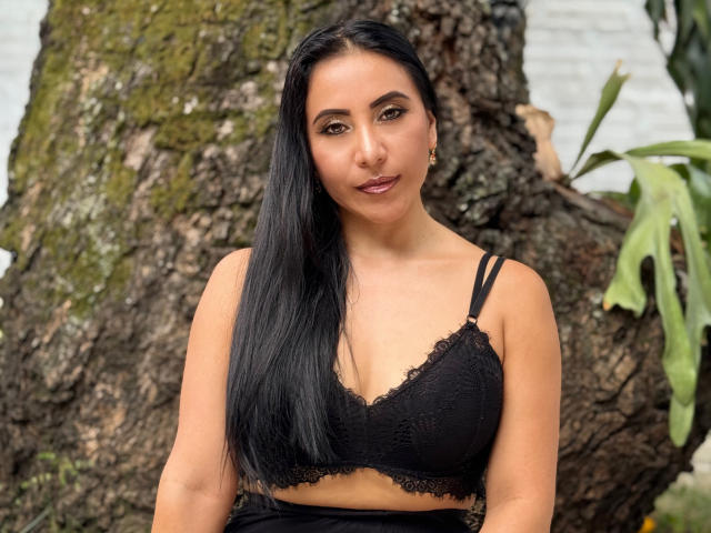VivienneWest, con lunghi capelli neri, occhi marroni e seno di media grandezza, indossa un reggiseno di pizzo nero e sta in piedi di fronte a un grande albero dalla corteccia ruvida. Ha un sorriso appena accennato..