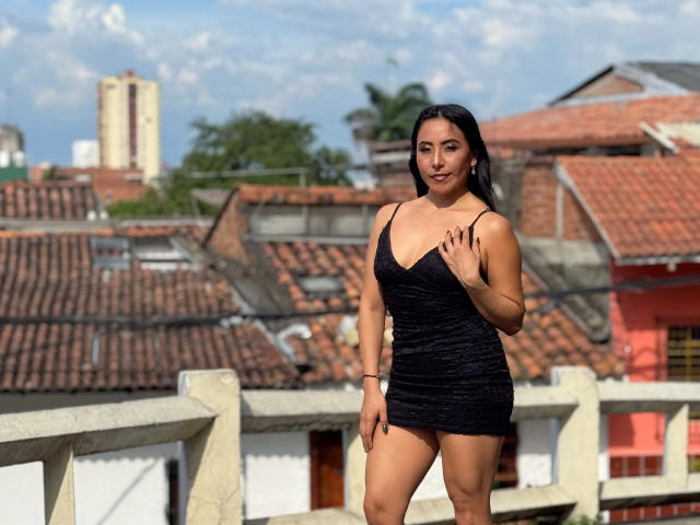 VivienneWest stands on a balcony in a black, sparkly mini-dress, showing her tits and ass. She's smiling, with long dark hair, and a hand on her chest.