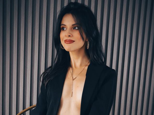 TaylaCooperk, avec de longs cheveux noirs, du rouge à lèvres, un blazer noir, un collier et des boucles d'oreilles dorées, souriant à la caméra, montrant une petite partie de sa poitrine nue..