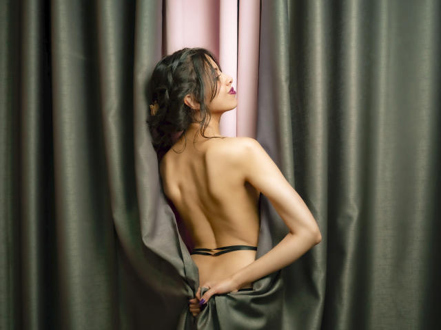 TaylaCooperk's bare back, small black thong, dark hair in a loose bun, facing a pink curtain, holding green curtains with her right hand, eyes closed, lips painted dark.