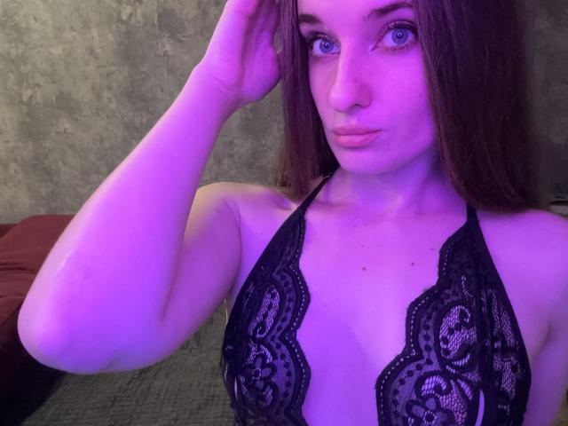 KariMillie with purple-lit skin, blue eyes, and brown hair, wearing a black lace halter top, showing her tits, hand in hair, looking at camera with pouty lips.