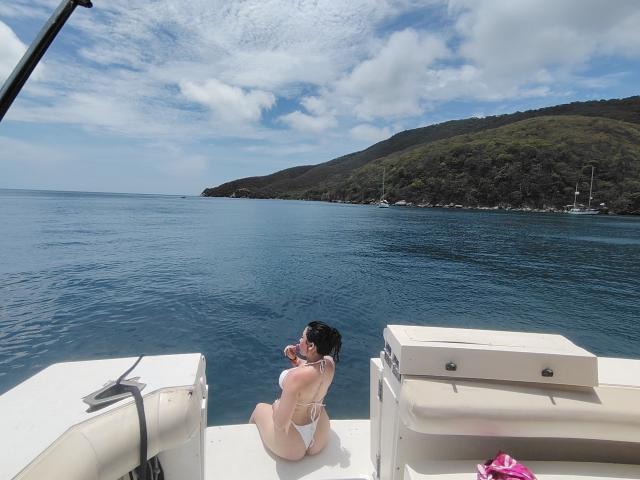 LunaBellaHotty in white bikini, sitting on boat with big ass, looking back at camera, holding phone, blue ocean, green hill in background, sunny day, boat seats on left.